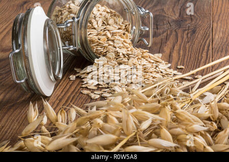 Rolled oats and dry ears on wood table. Common oat. Avena sativa. Beautiful close-up of a lying glass jar. Spilled flakes, dry cereal spikes, grains. Stock Photo