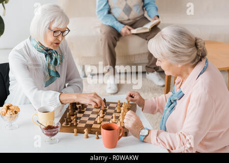 senior women playing chess and drinking coffee while man reading book on sofa Stock Photo