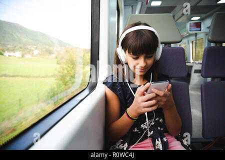 Happy little girl with headphones using her smart phone while travelling by train Stock Photo