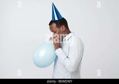Funny young african guy inflating the balloon wearing blue party hat. Stock Photo