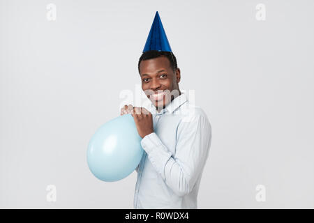 Funny young african guy inflating the balloon wearing blue party hat. Stock Photo