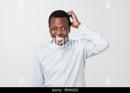 Confused african man scratching head while looking at camera and standing over gray background. Stock Photo