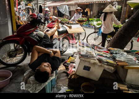 A man resting at his shop with women wearing conical hats in the background in the Old Quarter of Hanoi, Vietnam. Stock Photo