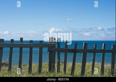 Pacific Ocean view from a grassy knoll behind a wooden barrier fence against a blue sky with puffy clouds Stock Photo
