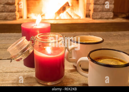 two white enameled mugs with hot tea, and candles , near cozy fireplace, in country house, winter vacation, horizontal. Stock Photo
