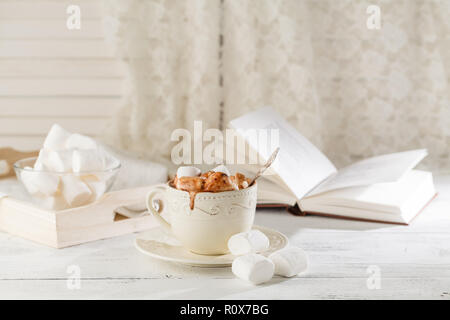 Mug of hot chocolate with marshmallows, on light wooden background with open book