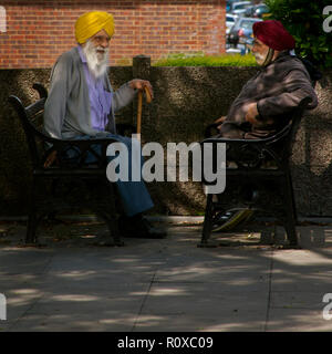 two elderly sikh man wearing yellow and red turbans sitting opposite each other on public benches Southall london Stock Photo