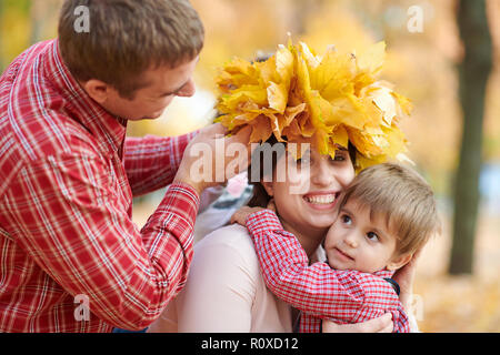 Father and son put yellow fallen leaves on mother head. Happy family is in autumn city park. Children and parents. They posing, smiling, playing and h Stock Photo