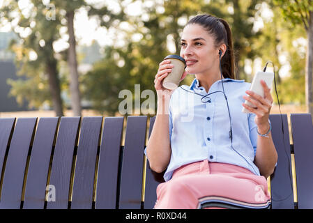 Young woman rests in autumn park on bench after a walk and listens to music on the phone at sunny day Stock Photo