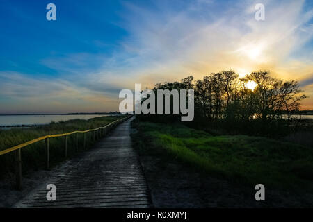 A lovely sunset view of a wooden walkway with handrail along the grass covered dunes & the calm sea. On the left side the sun shines through a... Stock Photo