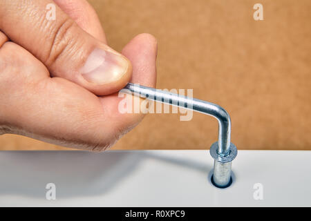 The handyman tightens the furniture connector bolts into the surface of wooden furniture with a hex wrench, close up, flat pack furniture assembly. Stock Photo