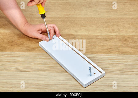 The furniture assembler tightens the connector bolts into a piece of wooden furniture with a screwdriver, with one hand he holds the furniture, the ot Stock Photo
