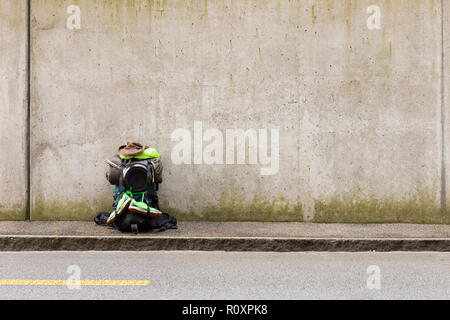 Hiking backpack with green towel, frying pan, sleeping bag, leather hat and green running shoes standing on a roadside in front of a gray wall Stock Photo