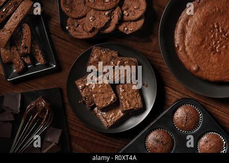 Flat Lay display of fresh baked chocolate desserts on black plates and a rustic wood table. Items are, Biscotti, Cake, Muffins, Brownies, and Cookies Stock Photo
