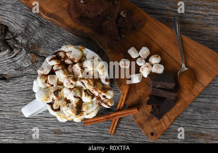 Overhead shot of a mug of hot cocoa with toasted marshmallows next to a cutting board with chocolate chunks cinnamon sticks and cookies. Stock Photo