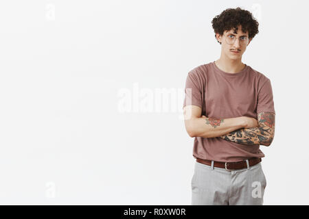 Attractive tattooed male feeling intense suspecting guy wants fool him around crossing hands on chest frowning looking serious and concerned at camera standing in glasses and stylish urban outfit Stock Photo
