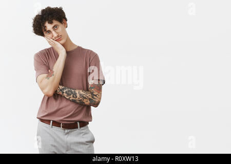 Guy hates sitting at home without action. Portrait of bored handsome and stylish urban young male with moustache, tattooed arm and curly hair leaning on palm indifferent and gloomy Stock Photo
