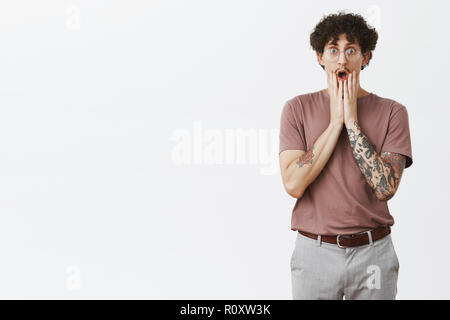Shocking news sweeping guy away from knees. Portrait of surprised astonished good-looking curly-haired male with tattoos on arms dropping jaw holding palms under mouth staring impressed at camera Stock Photo