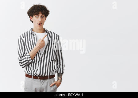 Indoor shot of impressed and amazed jewish curly-haired guy with moustache and tattoos dropping jaw and gasping from interest and excitement pointing at upper right corner intrigued Stock Photo