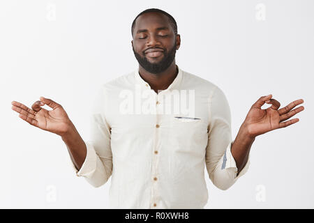 Peaceful and delighted African-american bearded male feeling nirvana smiling from pleasure as meditating with lotus pose and yoga orbs, close eyes releasing stress, feeling calm over gray wall Stock Photo