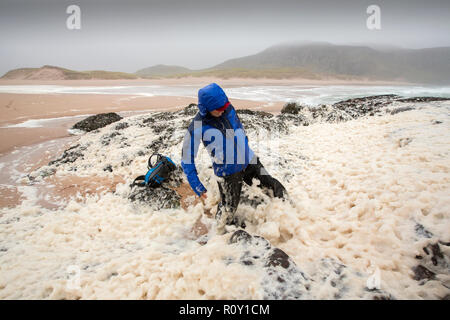 A hiker playing in spume or sea foam on the beach at Sandwood Bay during gale force winds, Sutherland, Scotland, UK. Stock Photo