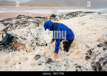 A hiker playing in spume or sea foam on the beach at Sandwood Bay during gale force winds, Sutherland, Scotland, UK. Stock Photo