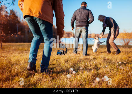 Videographer filming couple with dog in autumn park. Man using steadicam and camera to make footage. Video shoot Stock Photo