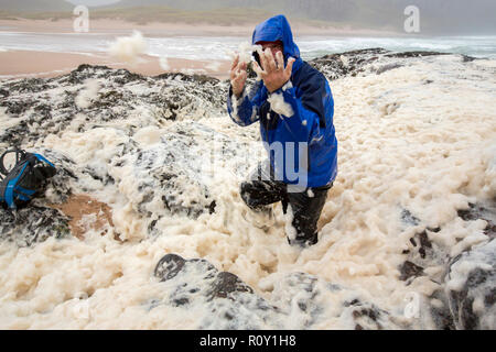 A hiker playing in spume or sea foam on the beach at Sandwood Bay during gale force winds, Sutherland, Scotland, UK. Stock Photo