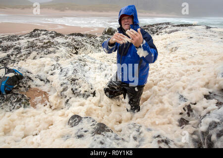 A hiker playing in spume or sea foam on the beach at Sandwood Bay during gale force winds, Sutherland, Scotland, UK. Stock Photo