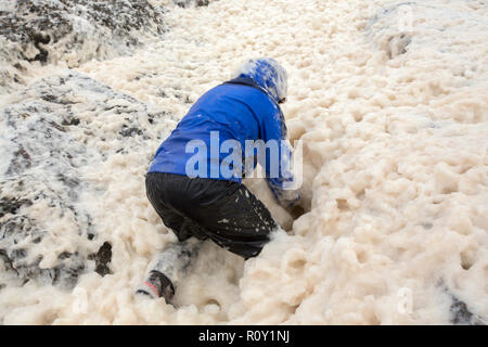 A hiker playing in spume or sea foam on the beach at Sandwood Bay during gale force winds, Sutherland, Scotland, UK. Stock Photo