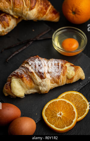 Croissants filled with italian custard surrounded with custard ingredients: orange slices, eggs and vanilla pods Stock Photo