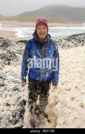 A hiker playing in spume or sea foam on the beach at Sandwood Bay during gale force winds, Sutherland, Scotland, UK. Stock Photo