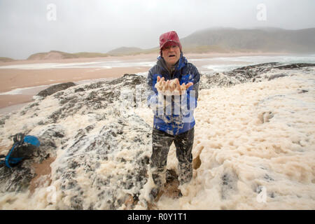 A hiker playing in spume or sea foam on the beach at Sandwood Bay during gale force winds, Sutherland, Scotland, UK. Stock Photo