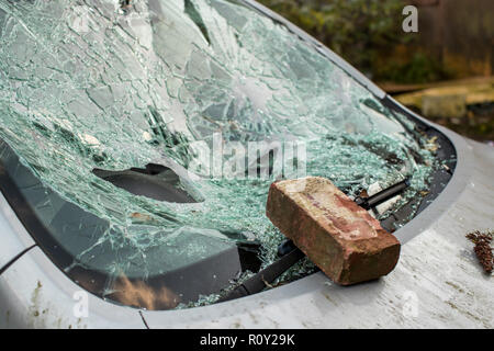 Brick sits on smashed windshield of a car with broken glass everywhere.  The car has been broken into and stolen. Car crime is a problem in United Kin Stock Photo