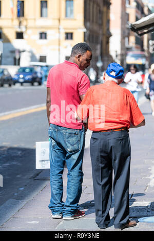 Rome, Italy - September 5, 2018: Back of African, black man helping senior old, elderly male, holding hand, assisting walking, carrying shopping bag,  Stock Photo