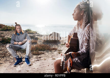 Blonde-haired man wearing blue sneakers listening to his woman playing guitar Stock Photo