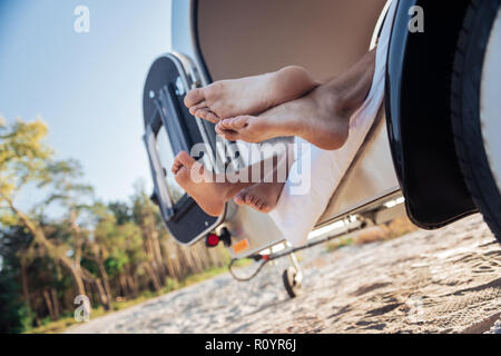 Couple of barefooted travelers lying in their compact trailer standing near beach Stock Photo