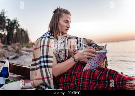 Loving cute couple with plaids on shoulders reading book after picnic Stock Photo