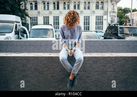 Unrecognizable young woman holding smart phone and looking aside sitting on parapet near european building and parking with cars. Stock Photo