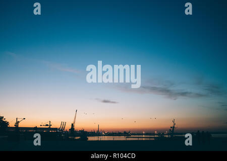 Silhouette industrial port against blue and orange sunset sky. Beautiful mood city landscape with urban lights and cargo cranes on sea. Stock Photo