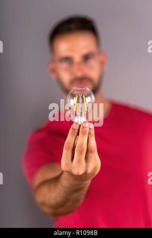 A young man in a red tshirt holding a lightbulb and standing in front of a grey background in the studio. Stock Photo