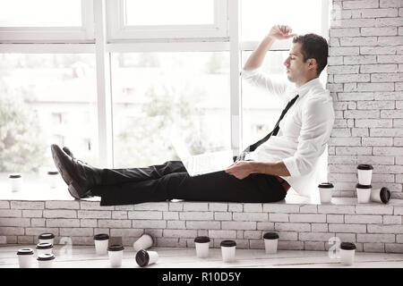 A young Manager sits on the windowsill in the office with a laptop and a bunch of coffee cups. Deadline. Stock Photo