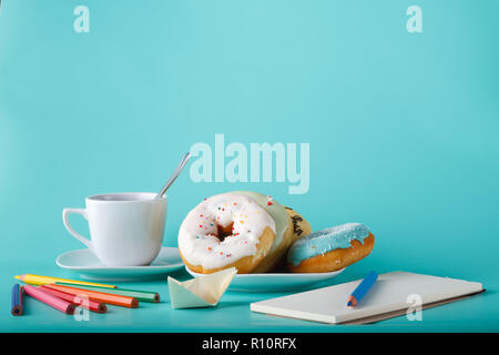 Four donuts on saucer with cup of tea. Aquamarine background with sketchbook and origami boat Stock Photo