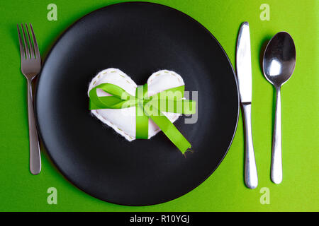 White heart on a plate on Valentine's Day. Elegant cutlery. Pantone greenery color theme. Green ribbon bow. Stock Photo