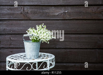 Bouquet of white cut flowers Lily of the valley (Convallaria majalis) in a zinc bucket shaped vase, outdoors on a white   fancy flower stand, brown wo Stock Photo