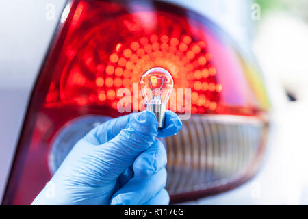 A hand in blue glove holding a car bulb, tail light in the background Stock Photo