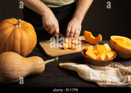 The cook slices the pumpkin for baking. On a black wooden table are whole pumpkins of different sizes. Stock Photo