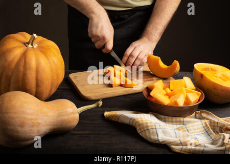 The cook slices the pumpkin for baking. On a black wooden table are whole pumpkins of different sizes. Stock Photo