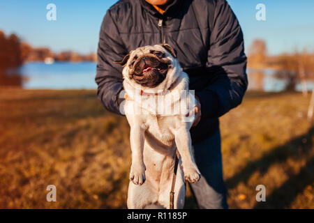 Master holding pug dog in autumn park by river. Man playing with pet.