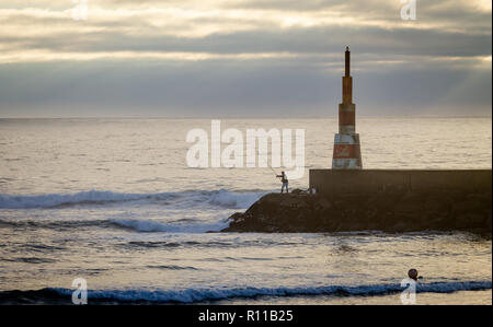A fisherman fishes near a beacon in the ocean, cloudy day.
